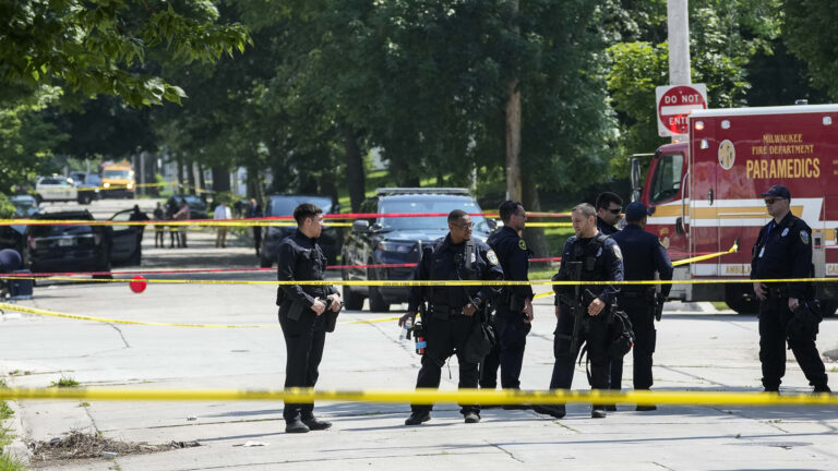 Multiple police officers stand in a street near an intersection surrounded by multiple placements of crime scene tape, with an ambulance and police vehicle parked behind them, and additional vehicles parked on a tree-lined street in the background.