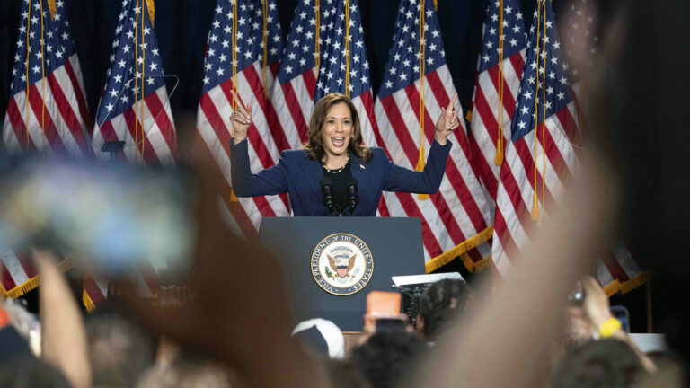 Kamala Harris gestures with both index fingers pointing upwards while standing behind a podium with a pair of microphones mounted to its top and the Seal of the Vice President of the United States on its front, with a row of U.S. flags in the background, and out-of-focus arms, hands and a phone screen of audience members facing her in the foreground.