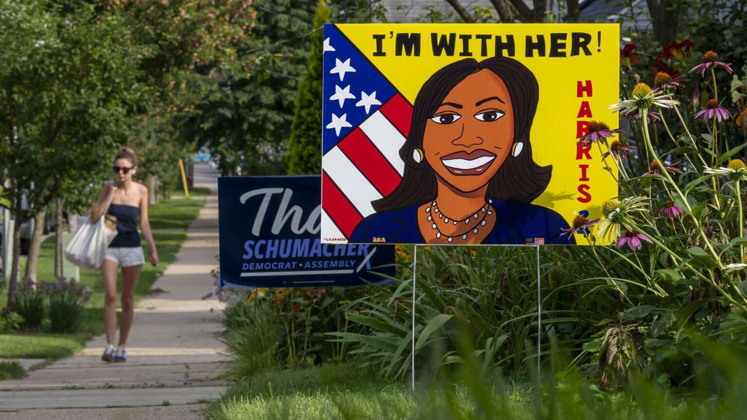 A printed yard sign showing a cartoon-style illustration of Kamala Harris in front of a U.S. flag with the words Harris and I'm With Her! stands on a lawn in front of another sign and garden plants, with a pedestrian walking along a tree-lined sidewalk in the background.