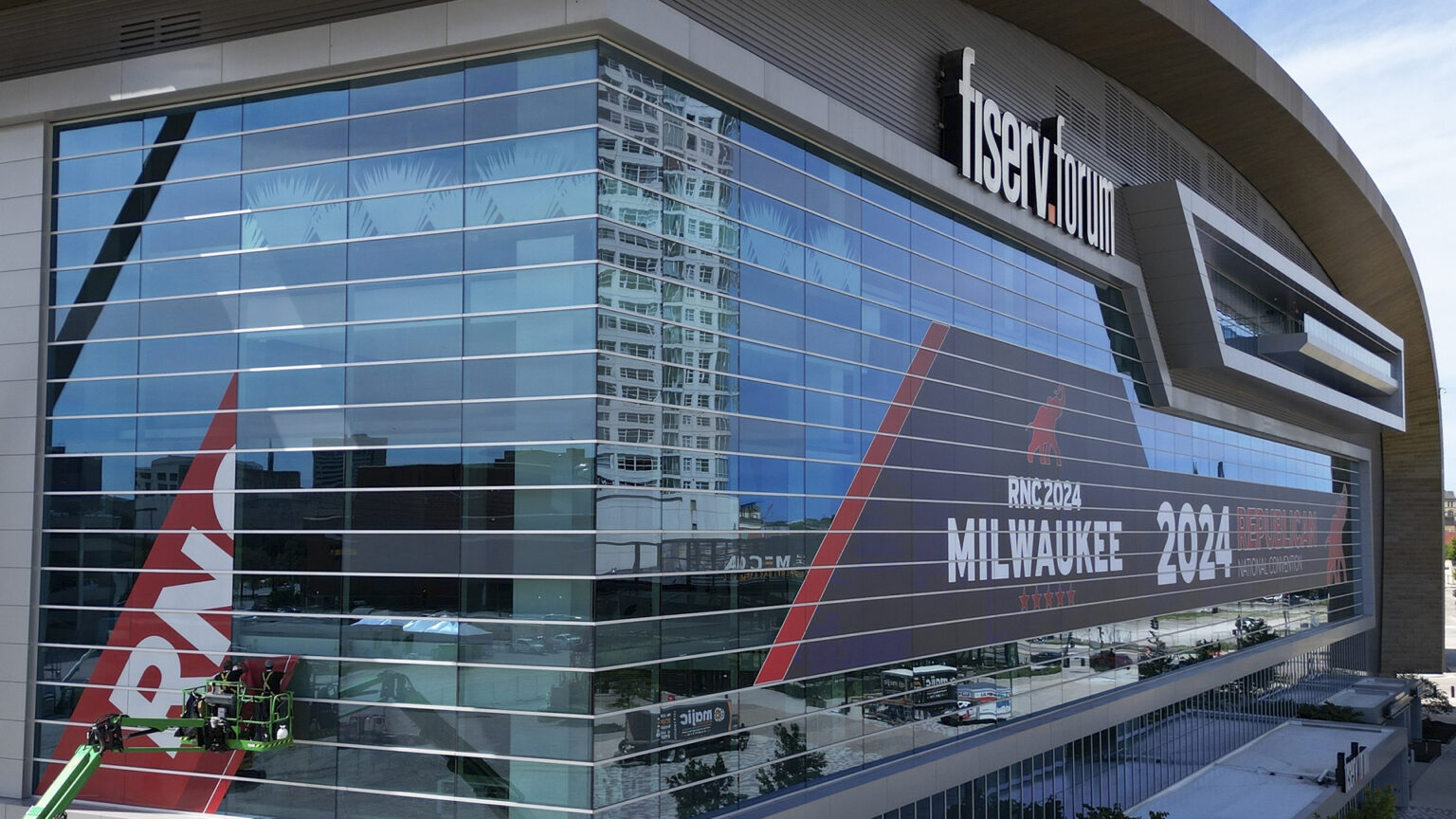 People standing on a boom lift apply decal material to a curtain glass wall of a large, multi-story arena with a curved roof, with a graphic on another face of the building reading RNC 2024 Milwaukee next to a stylized illustration of an elephant, with a an illuminated sign near the top reading fiserv.forum.