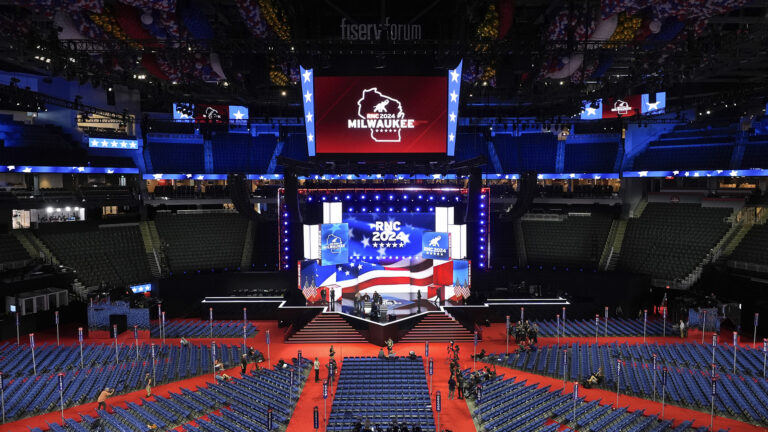 Multiple stadium screens show the RNC 2024 Milwaukee logo with an outline of Wisconsin and a stylized elephant illustration inside an arena with rows of permanent seats around the perimeter and a radial array of temporary seats arranged on the floor, with numerous balloons of different sizes contained behind temporary barriers under the high ceiling.