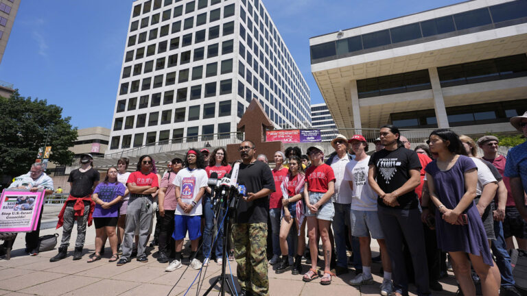 Omar Flores stands and speaks into multiple microphones with the flags of different media organization mounted to a stand, with dozens of other people standing behind on a plaza surrounded by multiple buildings of different heights.