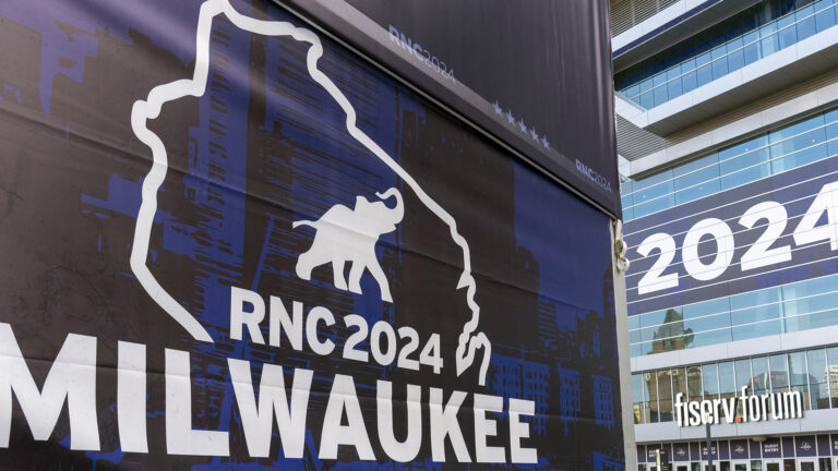 A graphic with the outline of the state of Wisconsin and an illustration of an elephant along with the words RNC 2024 and Milwaukee is displayed on a sign in front of a building with glass walls, which has another temporary sign that reads 2024 in part and permanent signage reading fiserv.forum above a row of doors.