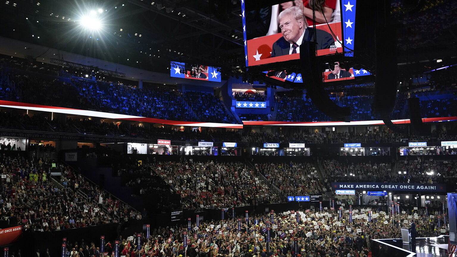 Multiple stadium screen mounted in the rafters of an arena shows Donald Trump while people in permanent seating in the stands and temporary seating on the floor watch a speaker on a stage.