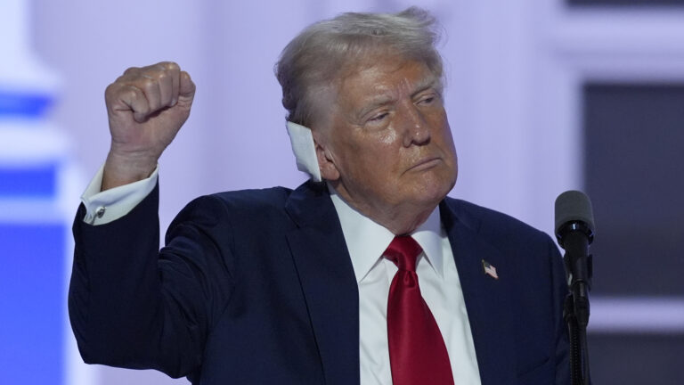 Donald Trump, with a bandage on his right ear, holds his right fist in the air while facing a microphone mounted on a stand.