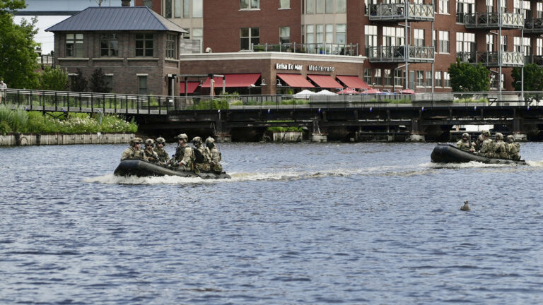 Two rigid hull inflatable boats filled with guardsmen wearing camouflage and military tactical equipment travel along a river, with a low bridge, a walkway and buildings in the background. 