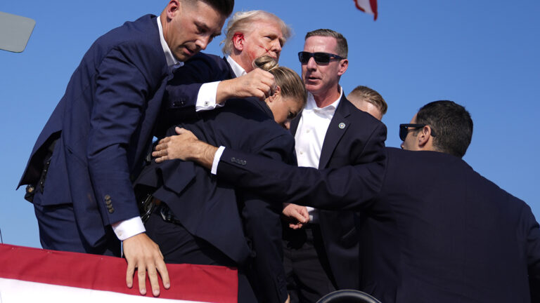 Five people surround and hold Donald Trump, who has blood on his right ear and cheek, while standing on a stage.