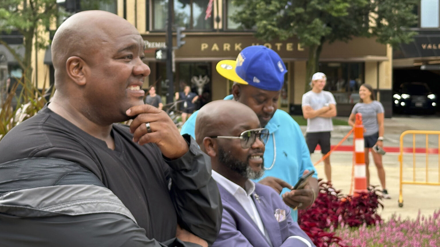 Michael Johnson smiles and holds his chin with his left hand while standing outside next to two other people, with pedestrians a building and trees in the background.