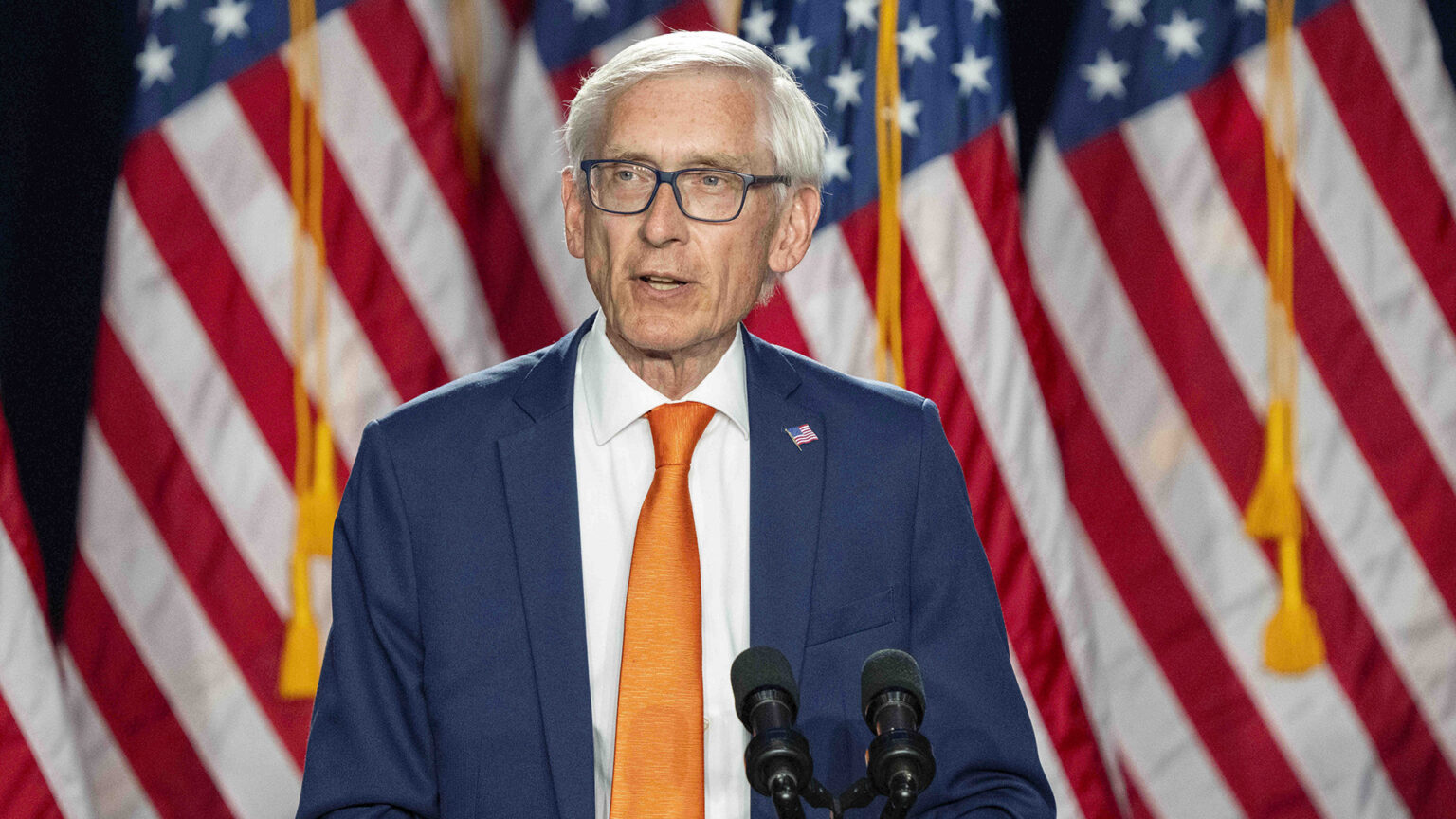 Tony Evers stands and speaks into a pair of microphones with a row of U.S. flags in the background.