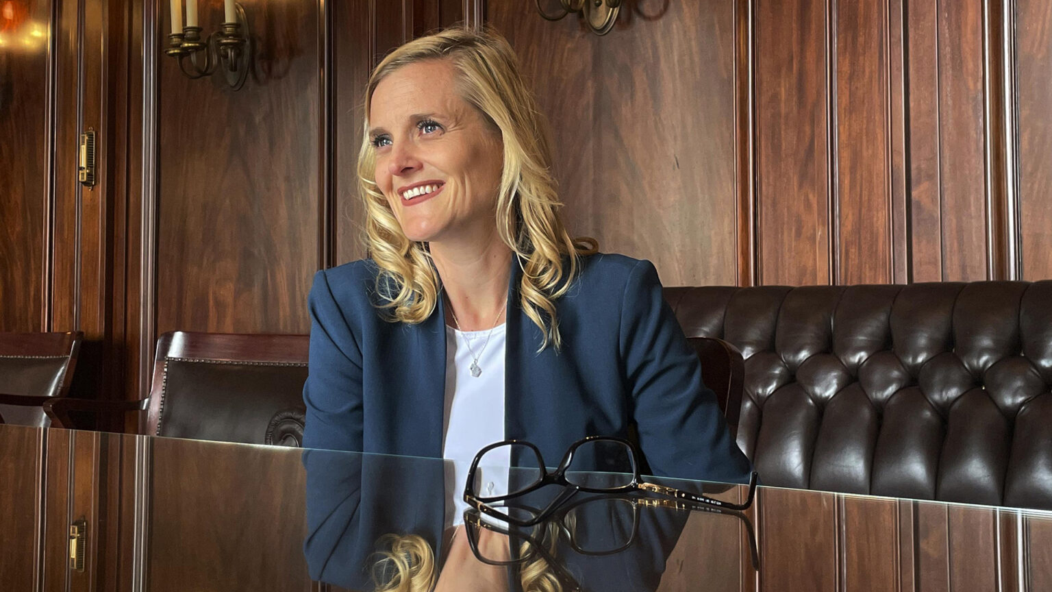 Sarah Godlewski smiles while sitting at a glass-topped table with a pair of glasses on its surface, in a room with wood and leather furniture, wood-paneled walls and electric wall sconces.