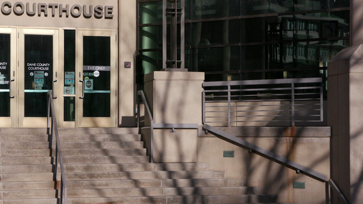 Letters reading Courthouse are affixed to a wall above a set of metal and glass doors at the top of concrete steps in front of a masonry and glass building.