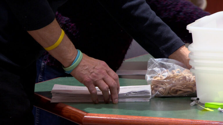 The hand of an election worker lifts an absentee ballot envelope from a stack on a table next to a plastic bag filled with rubber bands and a stack of plastic bins, with the arms of another election visible in the background.