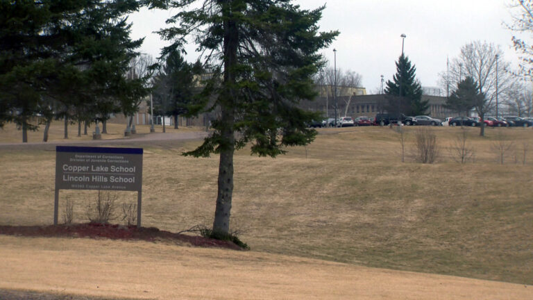 An entrance sign with the words Copper Lake School and Lincoln Hills School stands next to a tree and in front of a field with a tree-lined driveway leading to a parking lot filled with vehicles and multiple buildings surrounded by light poles.