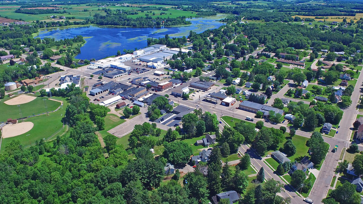 An aerial photo shows buildings, parks and trees situation among a grid of roads, with a pond behind the developed area and farm fields extending to the horizon.