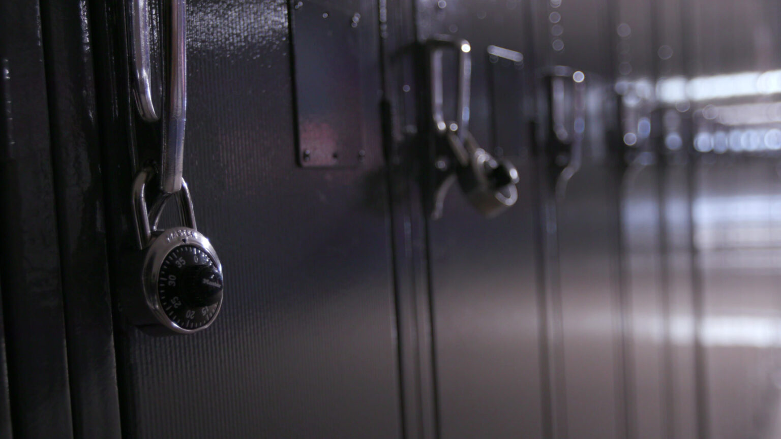 Combination locks secure two handles in a row of metal lockers in a hallway, with glare from fluorescent light fixtures reflecting off their painted surfaces.