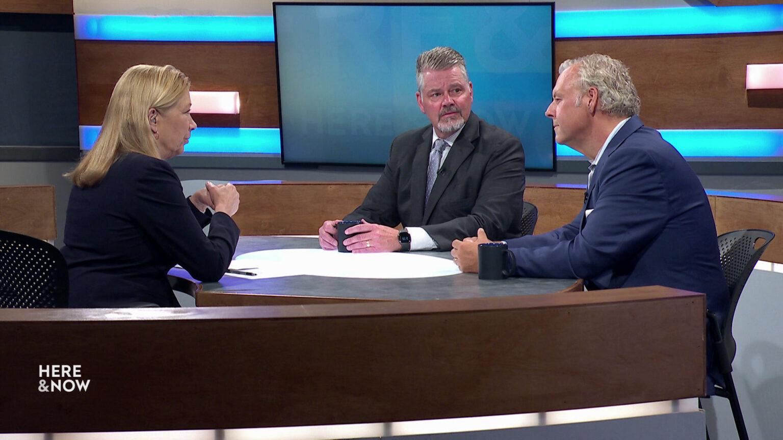 Bill McCoshen and Scot Ross sit next to each other and across from Frederica Freyberg at a desk on the Here & Now set.