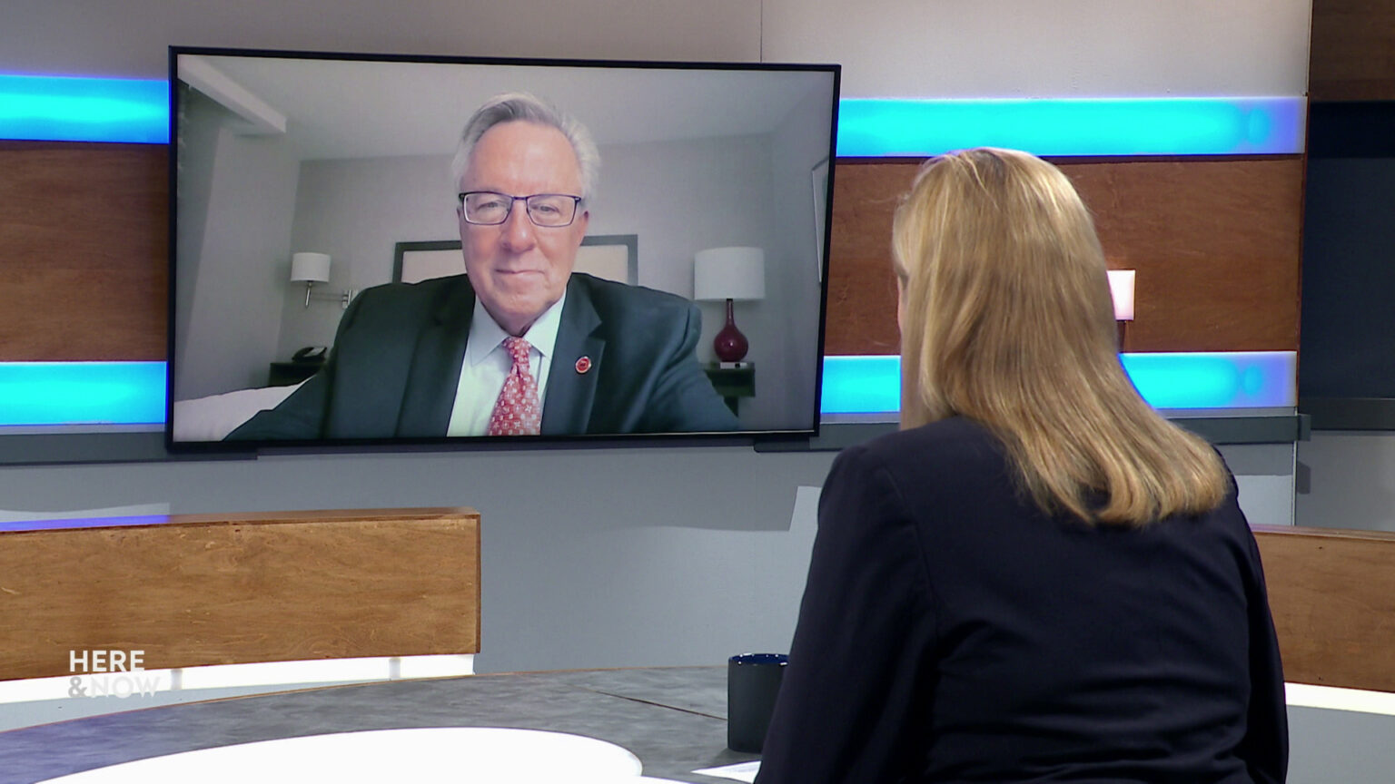 Frederica Freyberg sits at a desk on the Here & Now set and faces a video monitor showing an image of Brian Schimming.