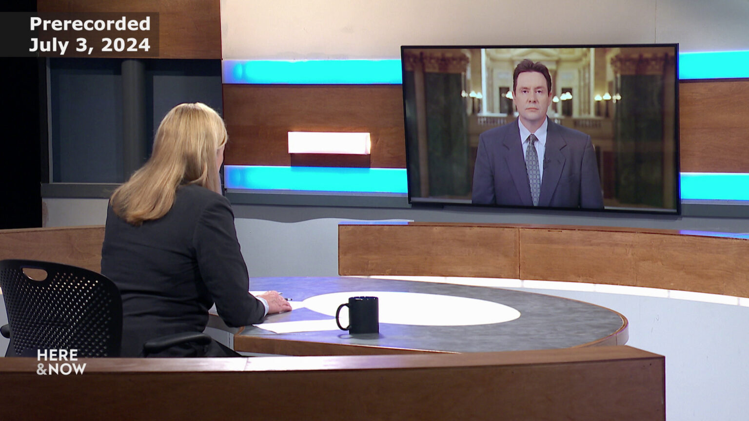 Frederica Freyberg sits at a desk on the Here & Now set and faces a video monitor showing an image of Zac Schultz.