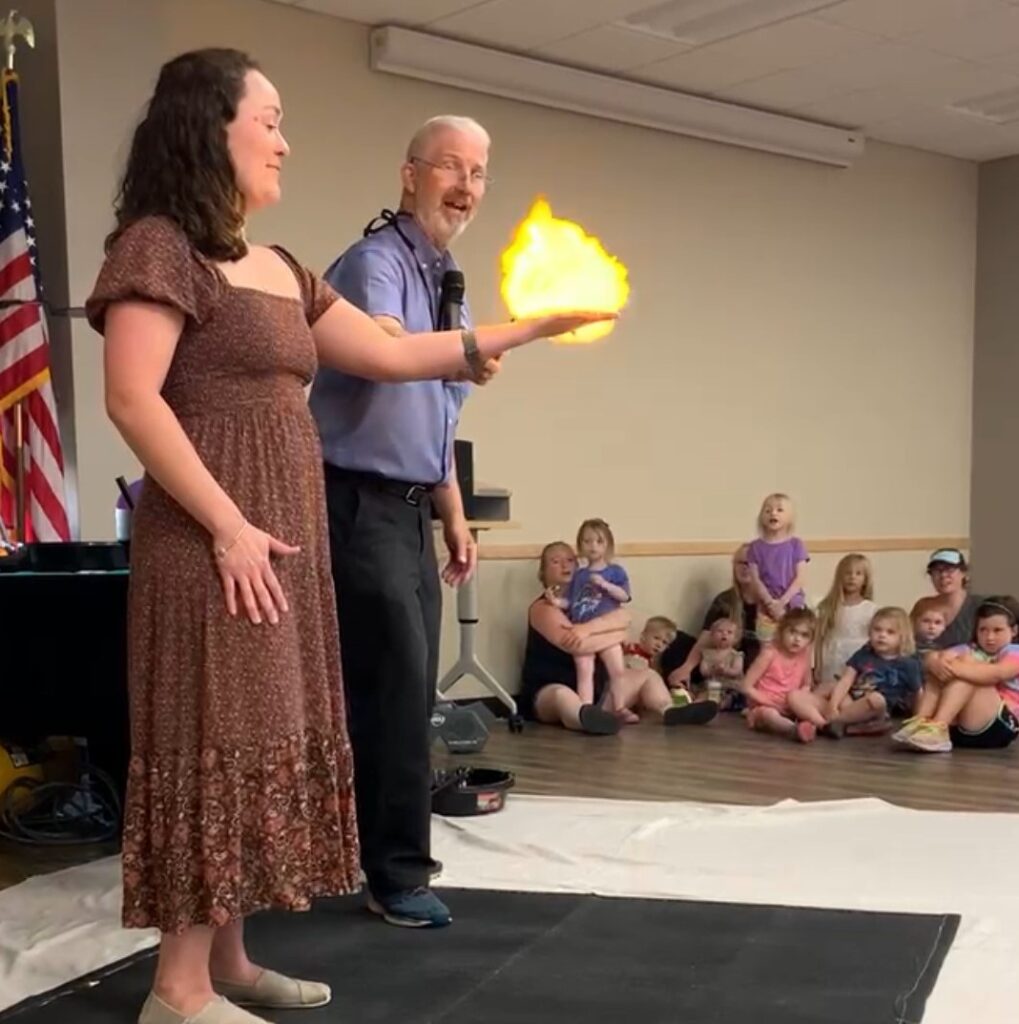 Two adults stand in front of an audience of families at a library. One adult is holding out her hand as a small burst of fire ignites. The audience looks excited.