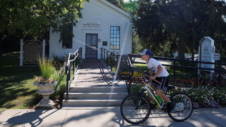 A child rides a bicycle on a sidewalk in front of steps leading to a brick-paved walkway in front of a small wood-sided building with a sign above its door reading Birthplace of the Republican Party and a Wisconsin State Historical Marker on one side, with trees in the lawn around the building.