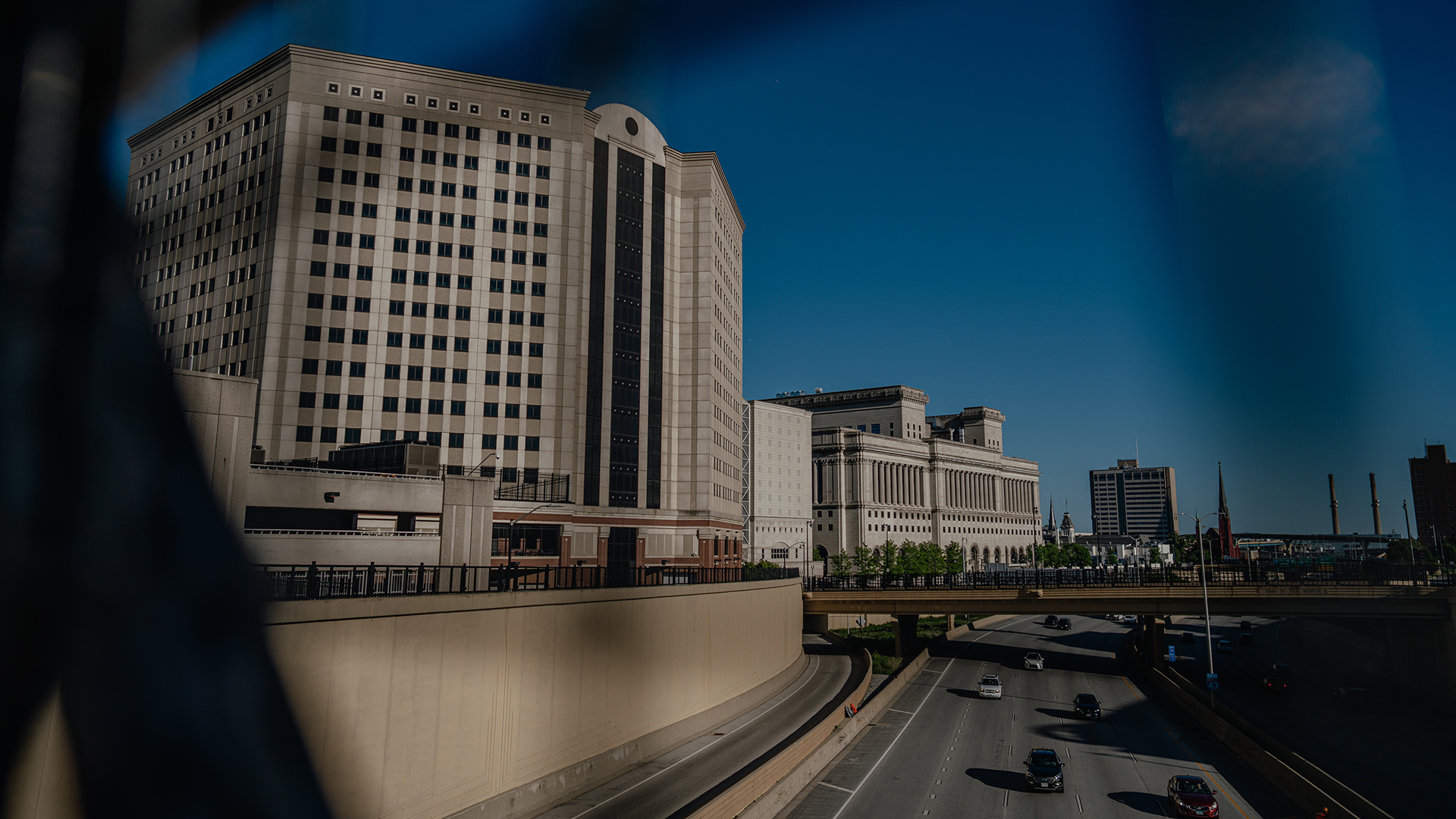 Cars drive on both sides of a below surface level divided highway, with a bridge, multiple masonry buildings, a church steeple and smokestacks in the background, with out-of-focus fencing obscuring portions of the view in the foreground.