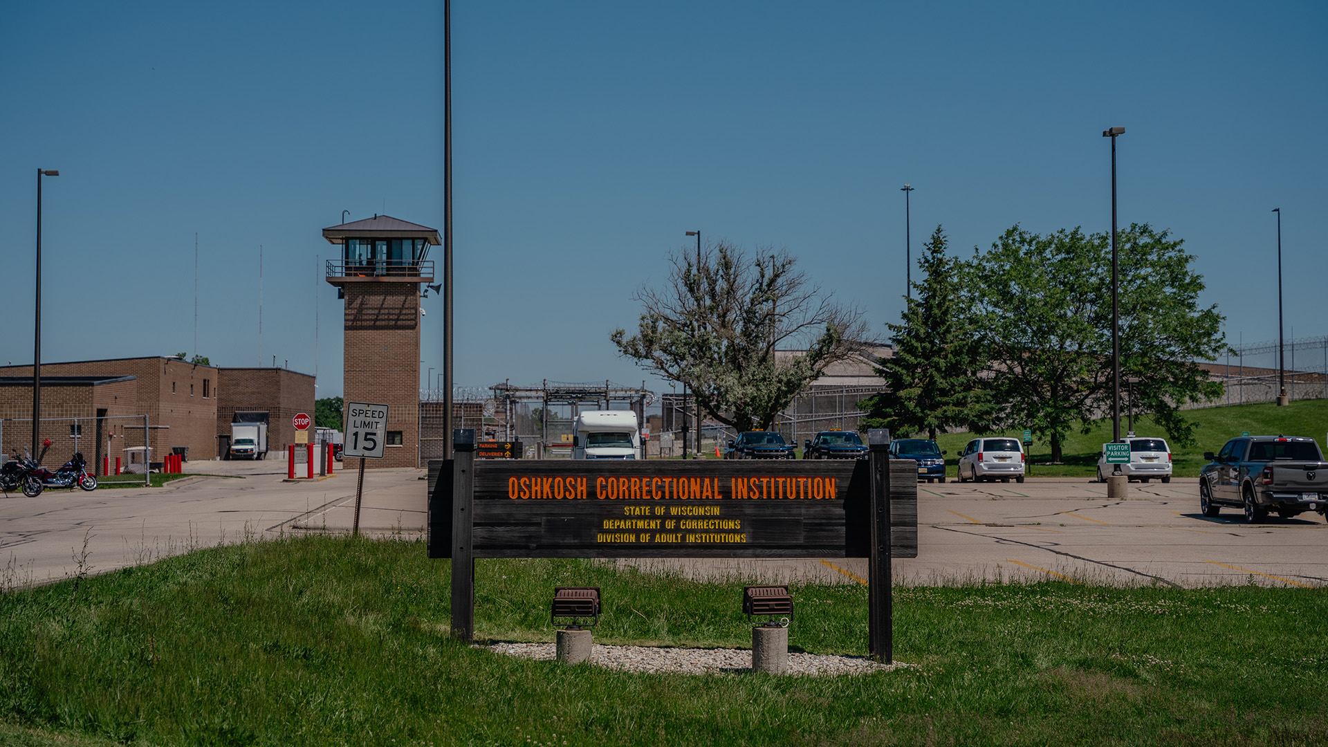 A wood sign with the words "Oshkosh Correctional Institution," and "State of Wisconsin Department of Corrections Division of Adult Institutions" painted on its surface stands on a lawn in front of a parking lot with multiple parked vehicles, with trees, a guard tower, light poles and fencing in the background.