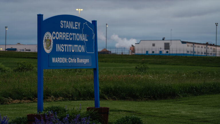 A sign showing Great Seal of the State of Wisconsin, an outline of the state and the words Stanley Correctional Institution stands in a field, with light poles and multiple buildings in the background surrounded by fencing.