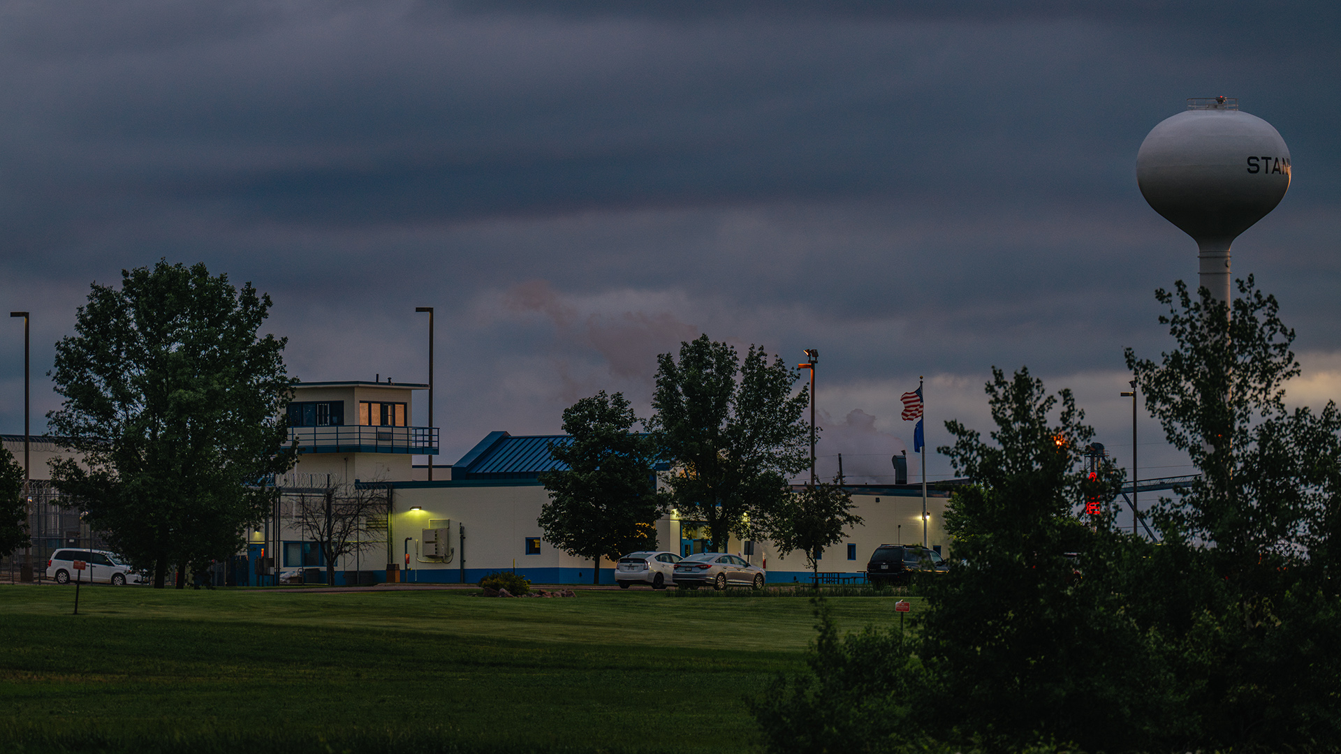 A water tower stands above a building surrounded by light poles, with a flagpole with the U.S. and Wisconsin flags, parked cars, trees and a lawn in the foreground.