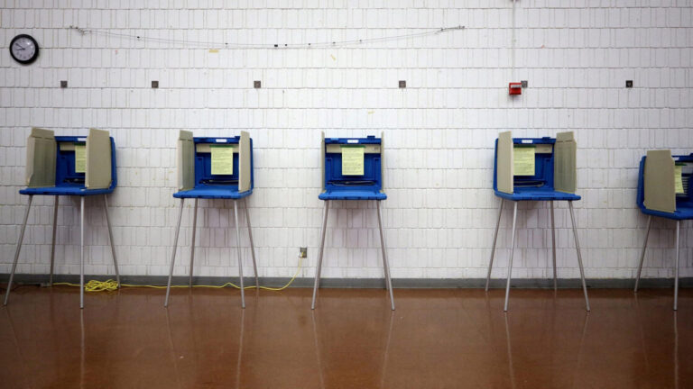 A row of collapsible voting privacy booths with a plastic base and walls and metal legs stands in a room with a linoleum floor and painted brick wall with a clock and fire alarm.