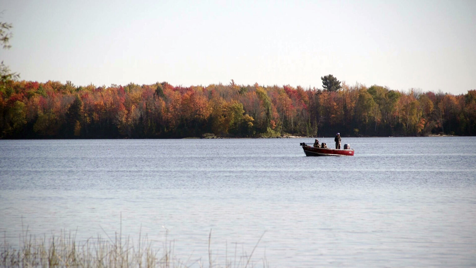 One person stands and two others site in an aluminum fishing boat with an outboard motor in a lake, with trees showing different colors of fall foliage on the opposite shore.