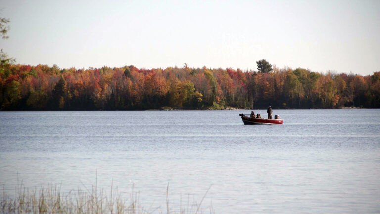 One person stands and two others site in an aluminum fishing boat with an outboard motor in a lake, with trees showing different colors of fall foliage on the opposite shore.