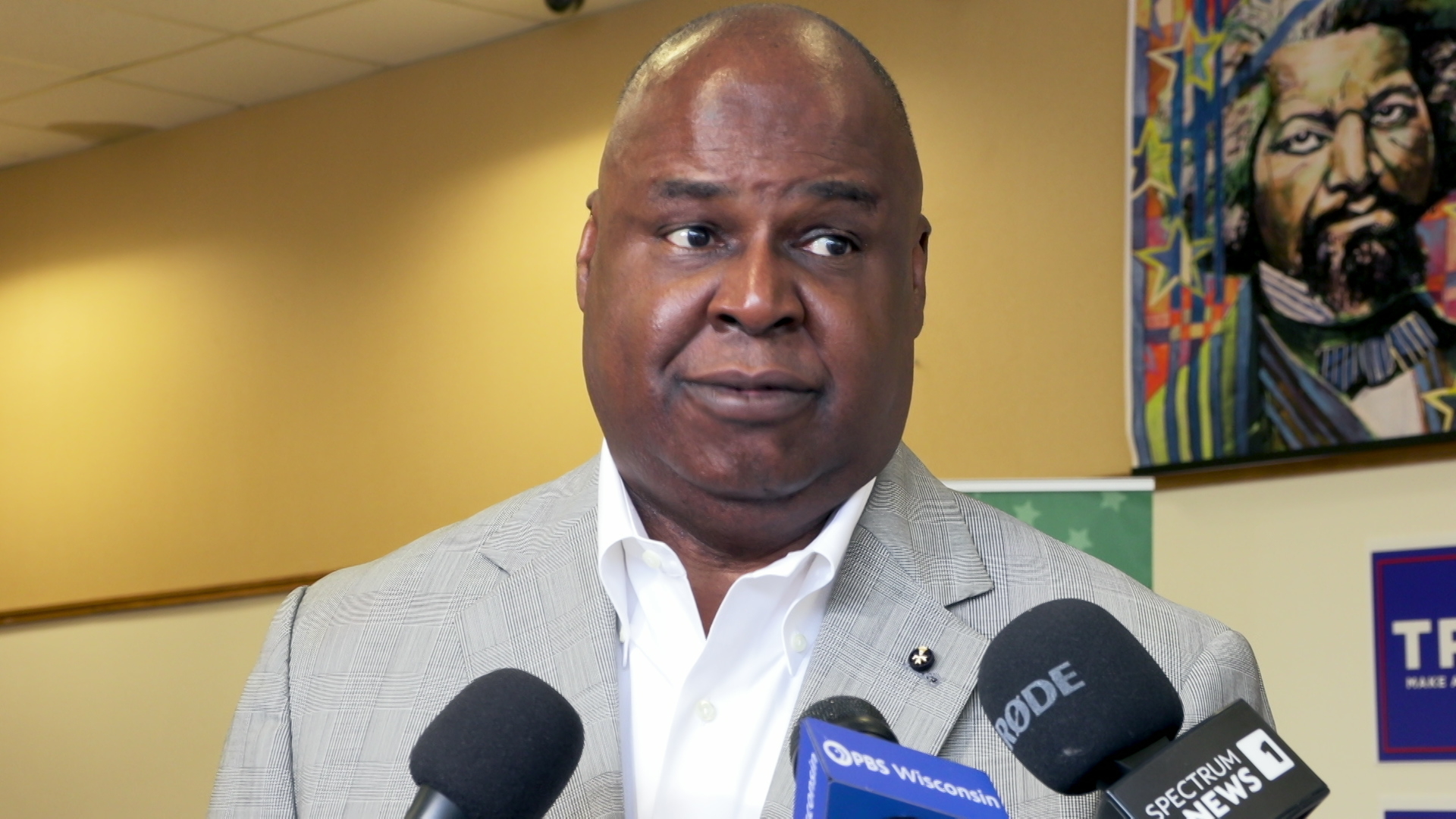 Gerard Randall stands and speaks into three microphones with flags that read "PBS Wisconsin" and "Spectrum News" inside with an illustration of Frederick Douglass in the background.