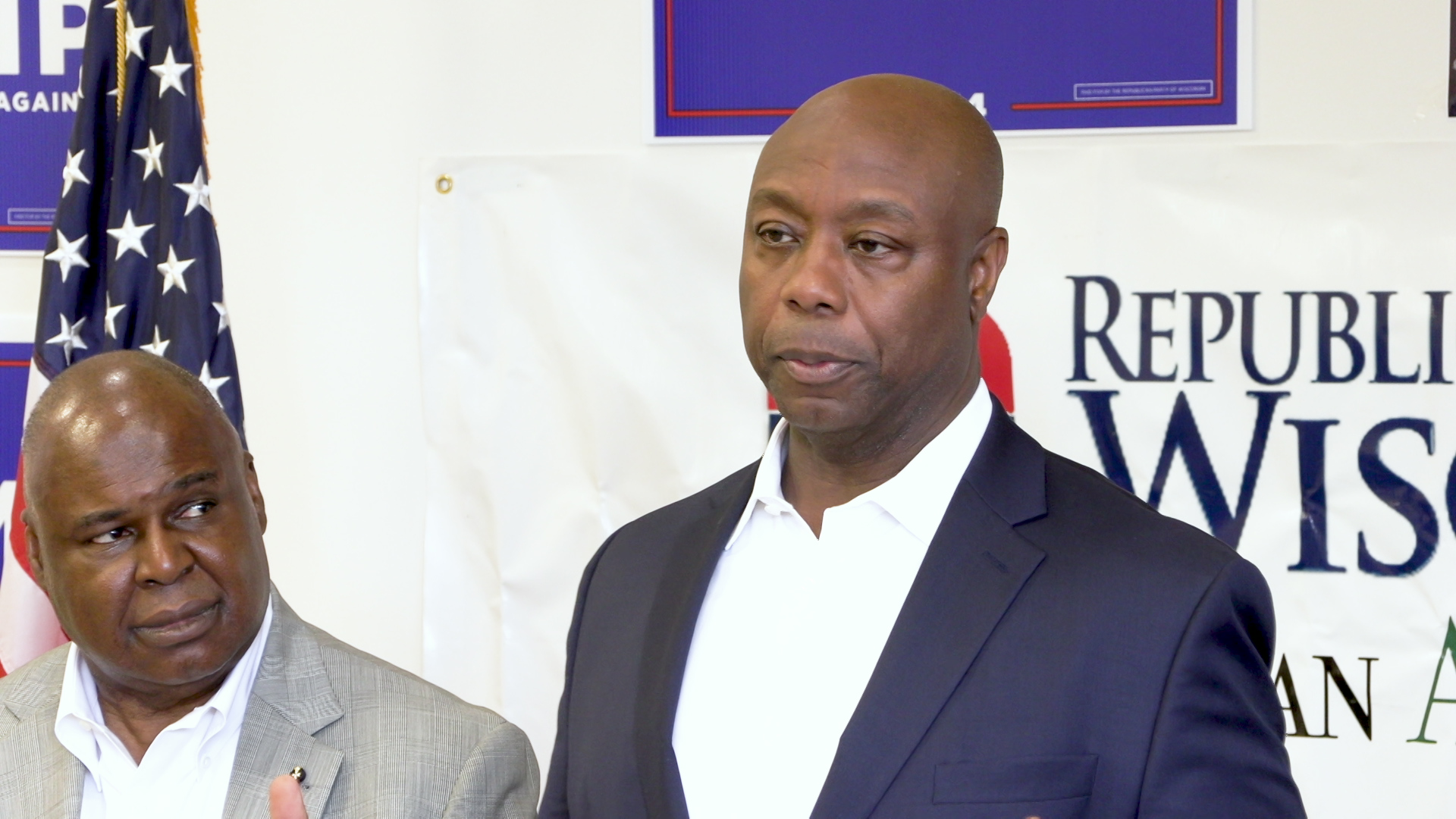 Tim Scott stands and speaks with another person, a U.S. flag and campaign signs in the background.