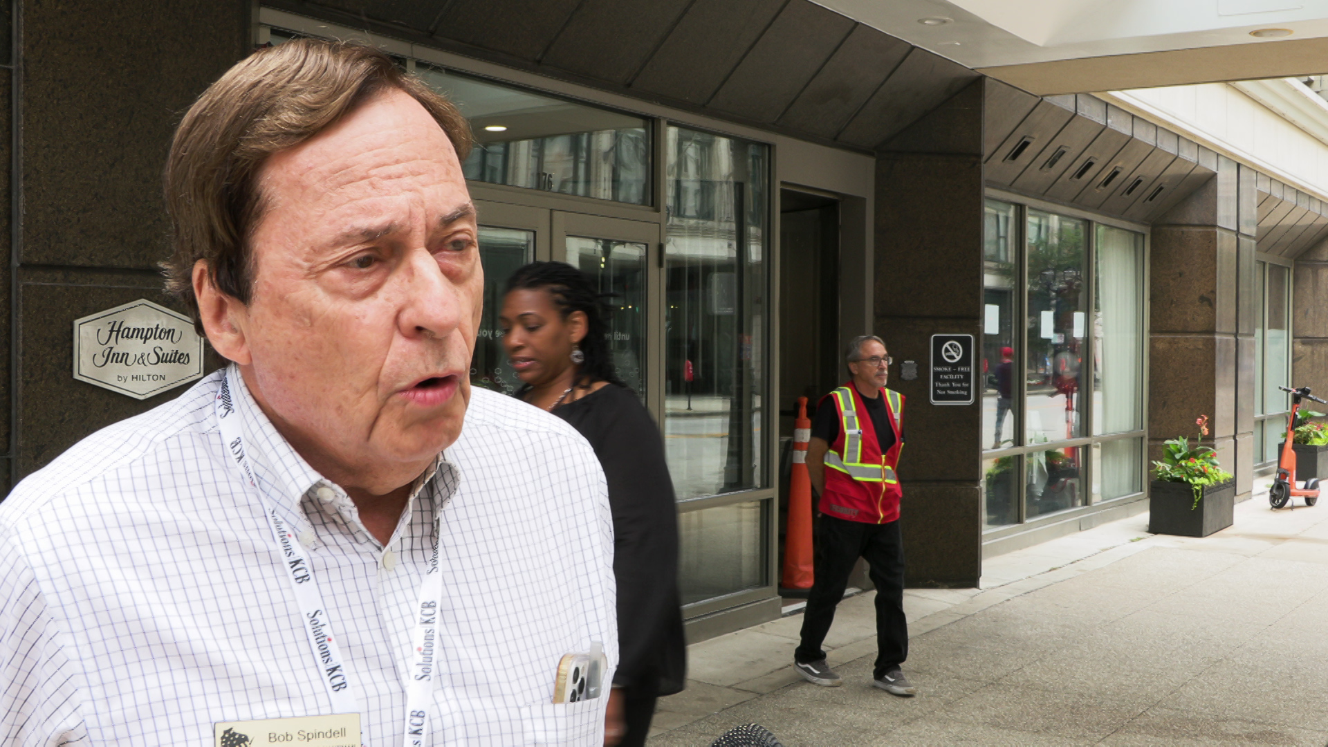 Bob Spindell speaks while standing on a sidewalk with people and a wall with glass doors and windows in the background.