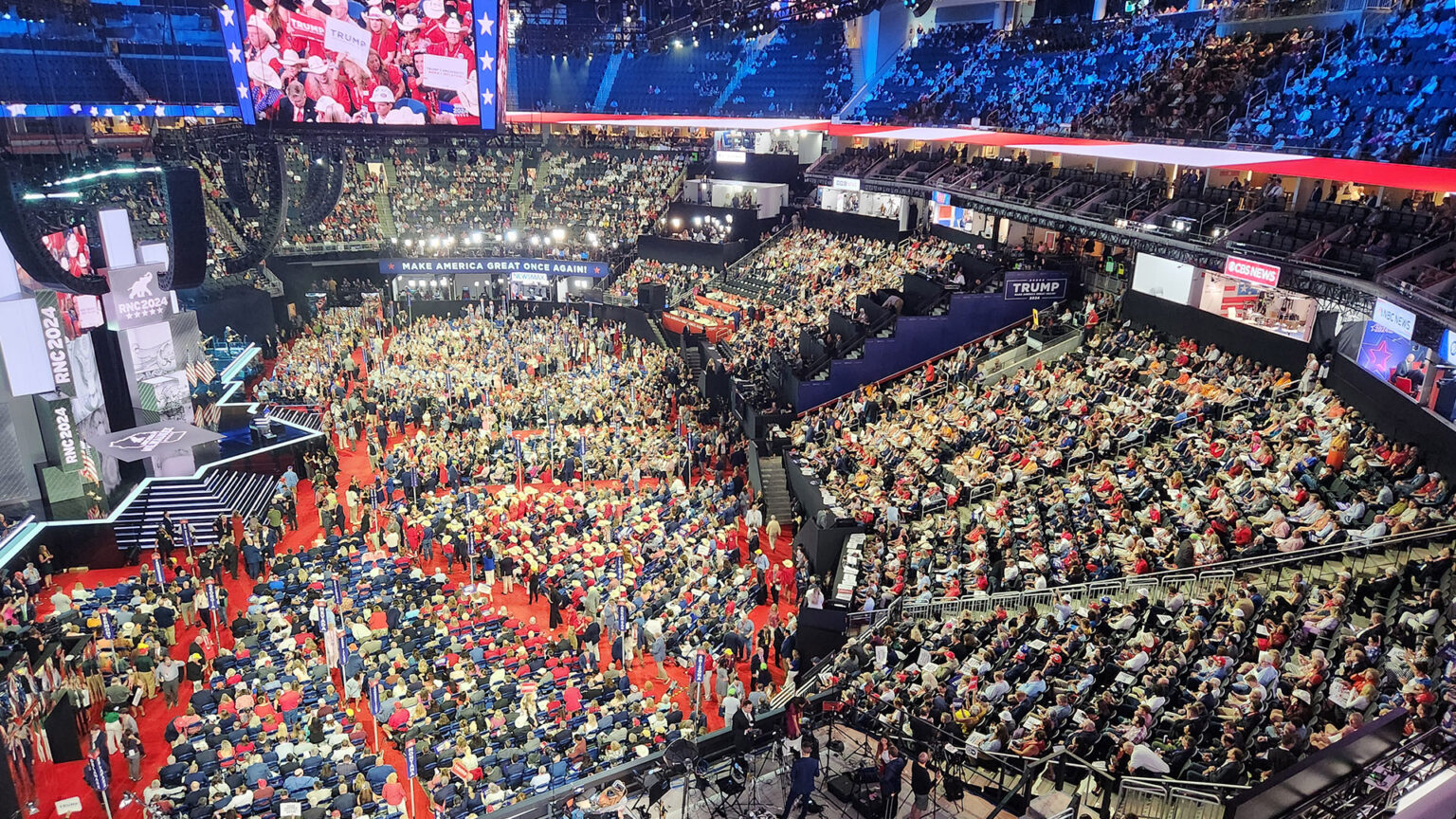 A four-sided stadium screen shows participants filling temporary seating on the floor and permanent seating in the stands of an arena, with a large stage with steps and a backdrop on one side of the space.