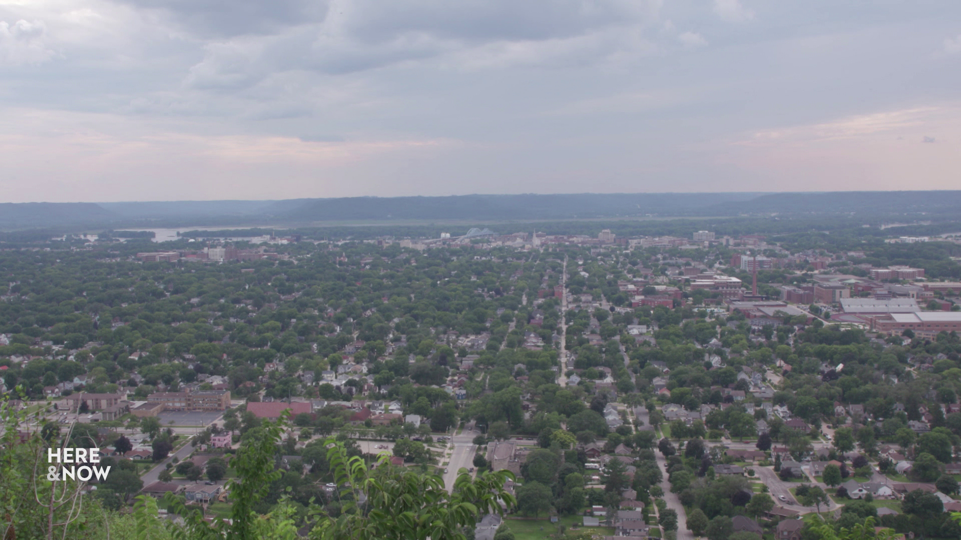 An aerial shot shows a grid of streets with trees and buildings extending to a river with bluffs on the far shore.