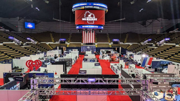 A four-sided stadium screen shows the RNC 2024 Milwaukee logo with an outline of Wisconsin and a stylized elephant illustration inside an arena with multiple rows of permanent seats around the perimeter and rows of temporary booths arranged on the floor.
