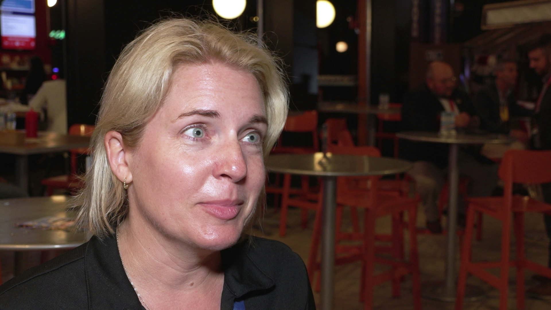 Marla Poytinger speaks inside a room with tables, chairs and people seated in the background.