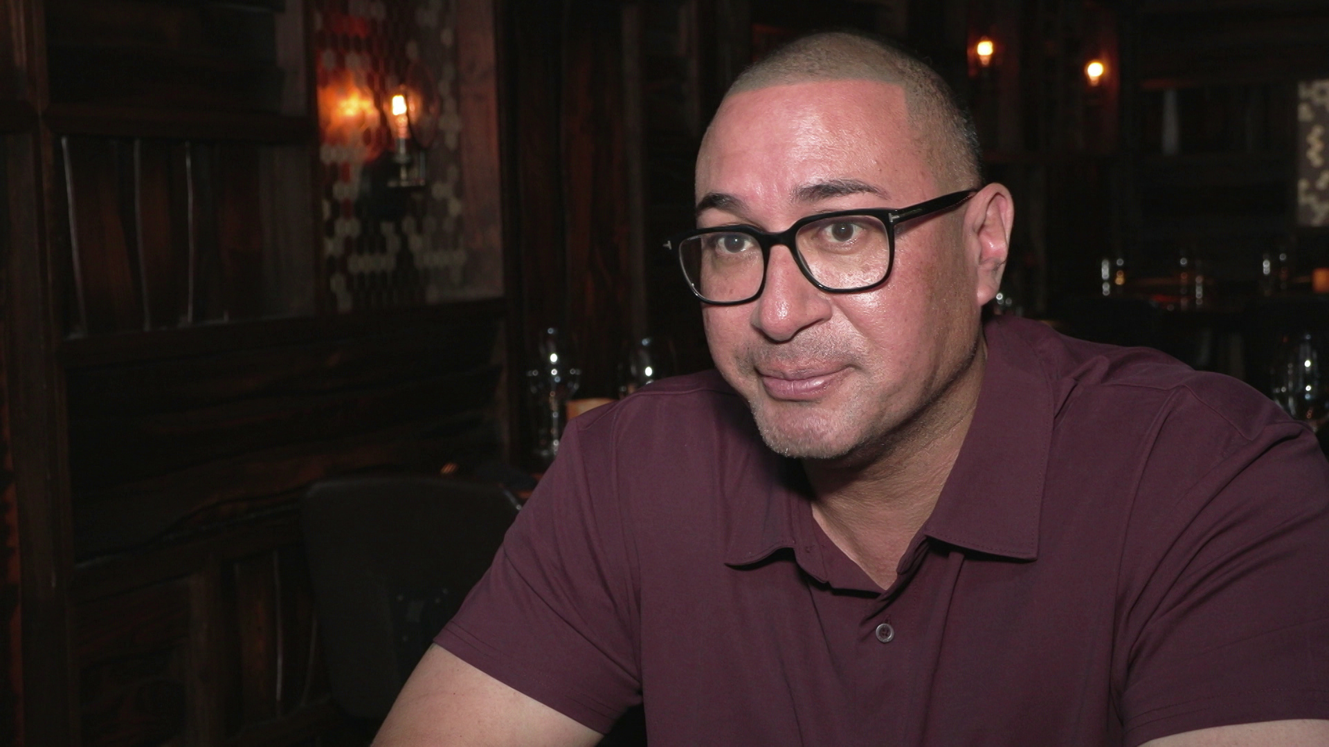 Omar Shaikh sits in a room with low lighting and wood paneled walls in the background.