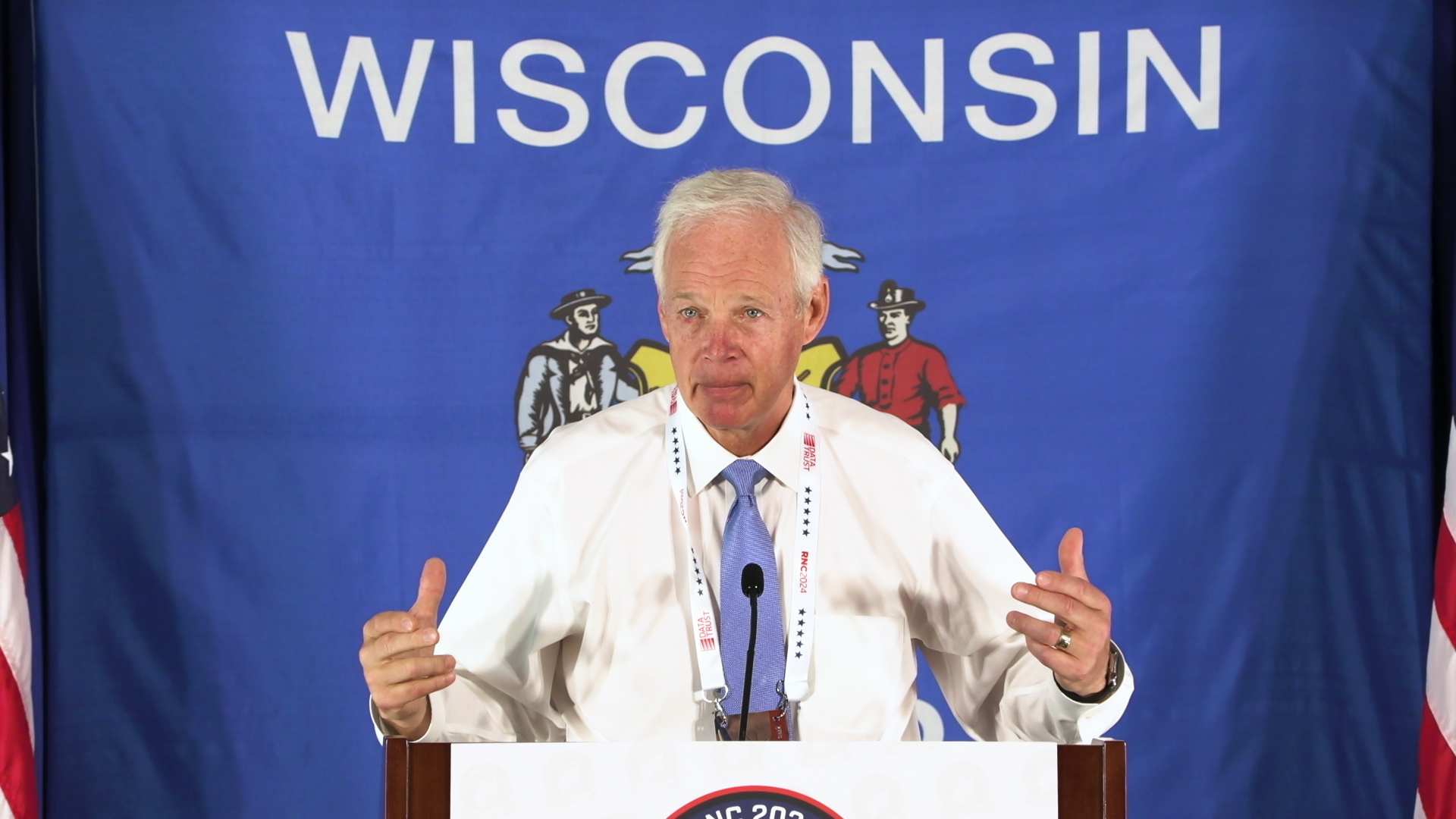 U.S. Sen. Ron Johnson stands behind a microphone on a podium with a Wisconsin state flag hanging up in the background.