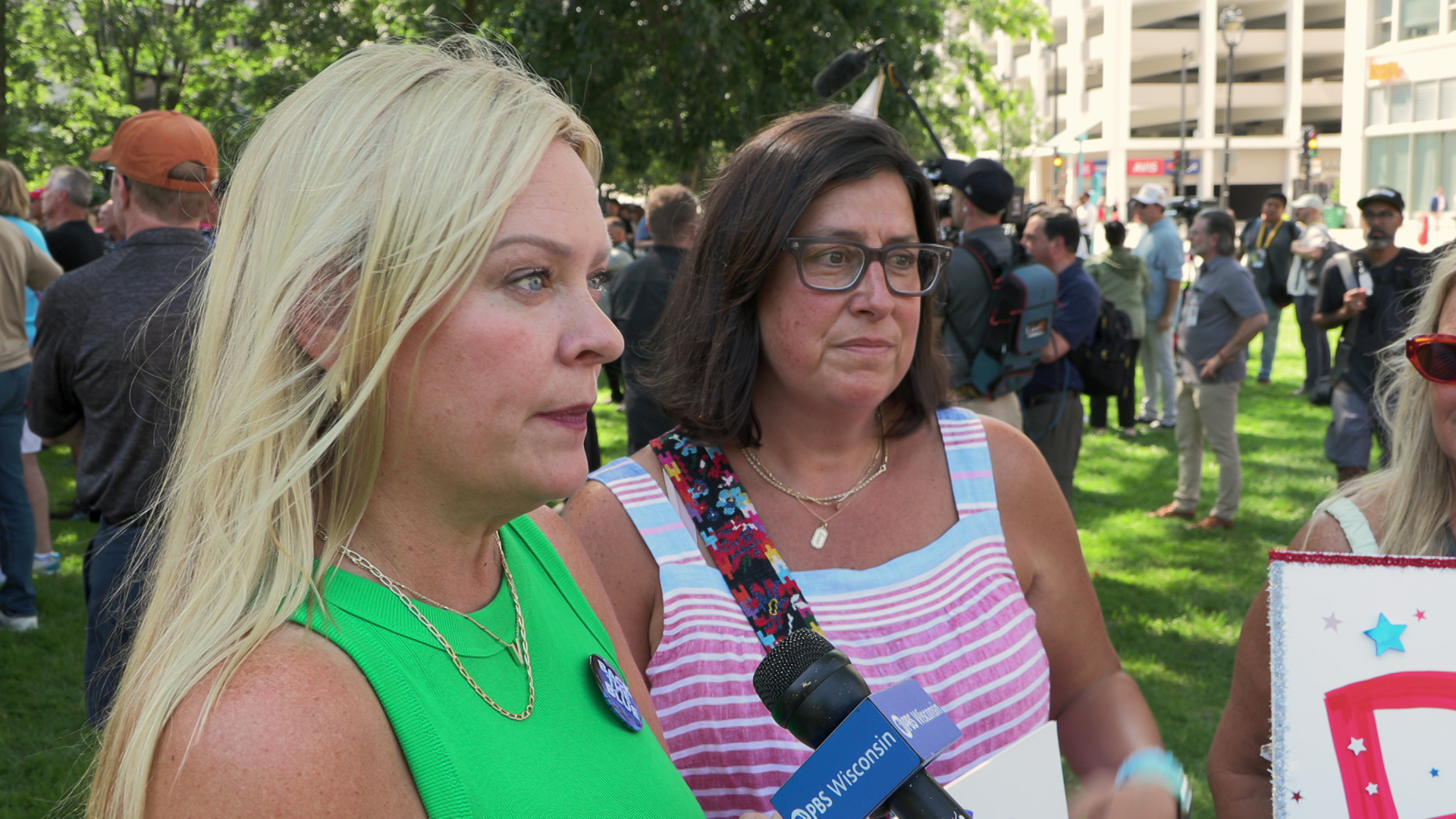 Valerie Mack speaks into a microphone with a flag reading "PBS Wisconsin" while standing outside next to another person, in front of other people with trees and buildings in the background.