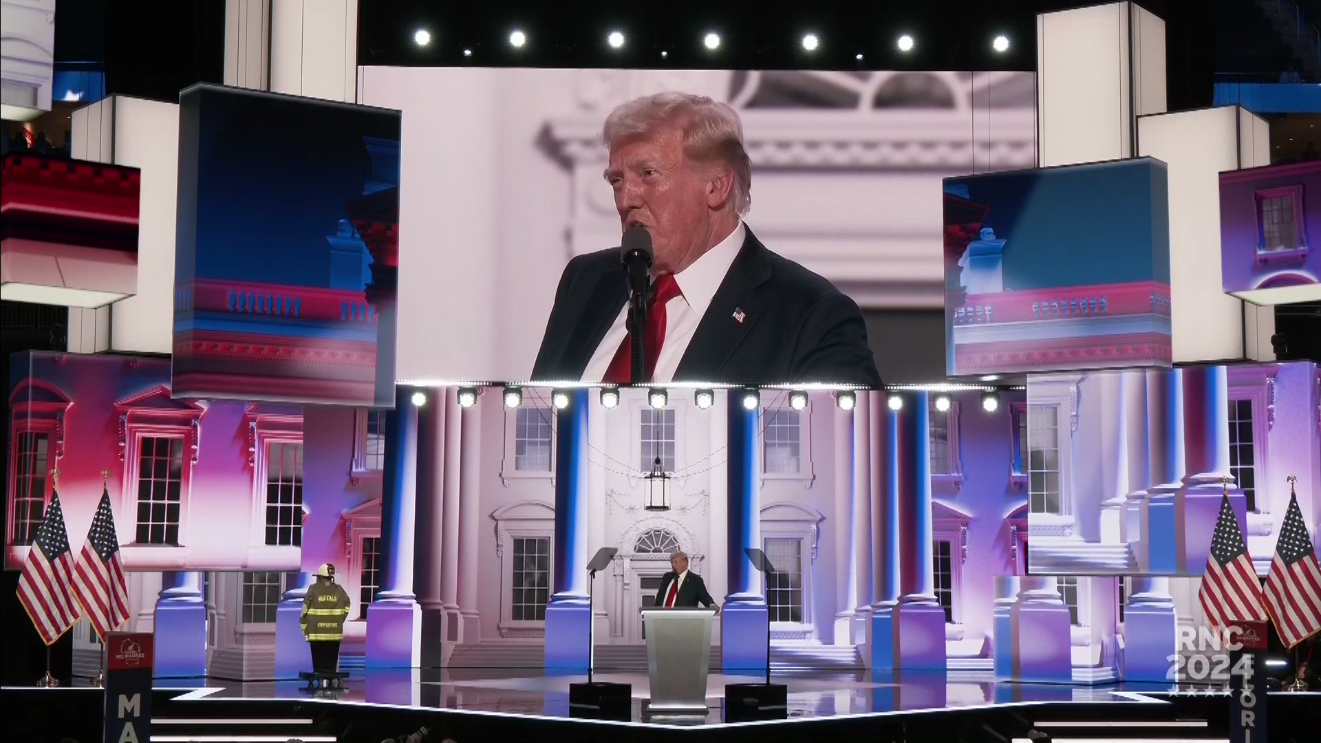 Donald Trump stands on stage behind a podium with a microphone and faces two teleprompter mirrors with a set featuring columns, the front face of the White House, U.S. flags a fire fighters uniform on a mannequin, and multiple screens behind him.