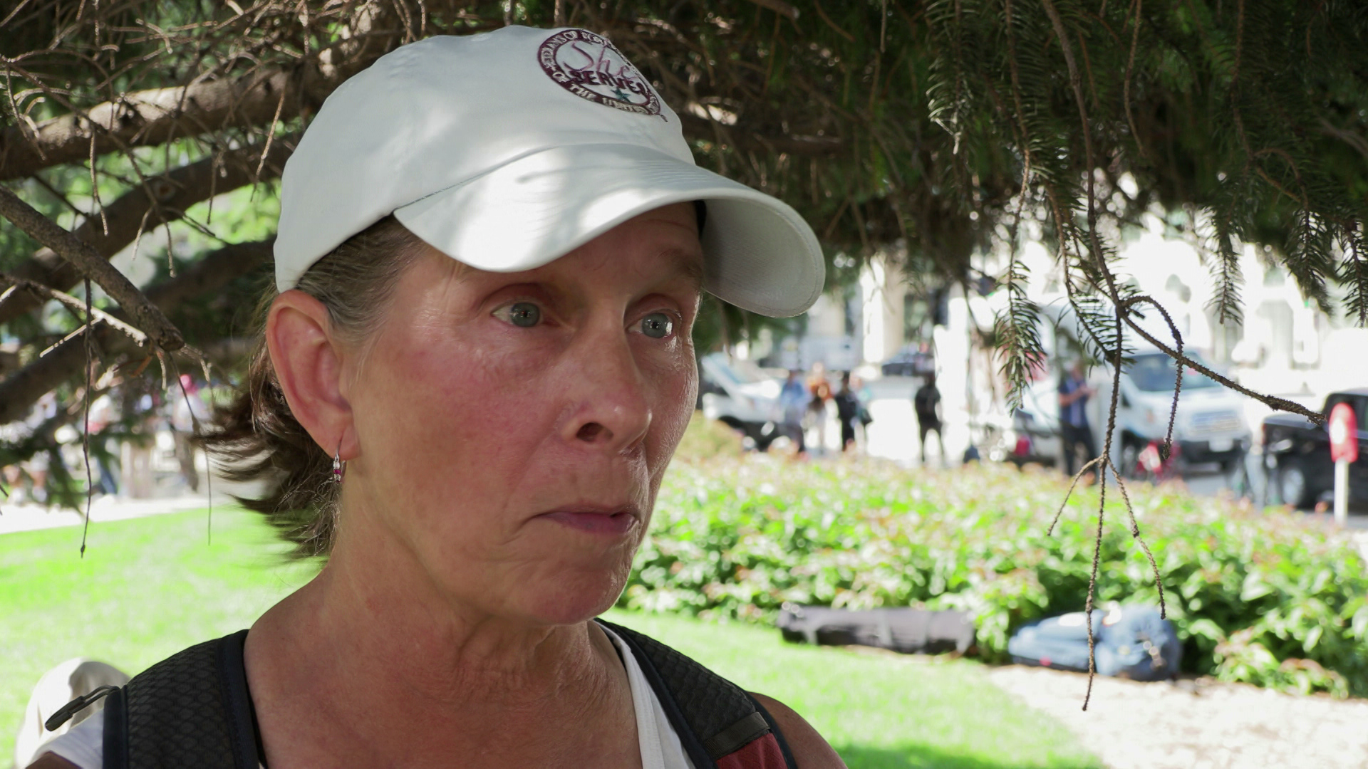 Ranay Blanford speaks while standing outside under a tree branch with a lawn, a flower garden and parked vehicles in the background.