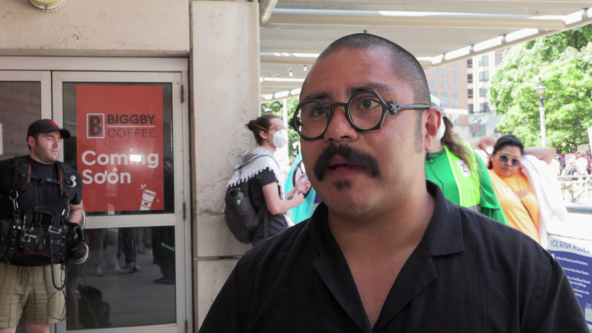 Omar Flores speaks while standing under metal roofing outside with protesters standing and walking behind him.