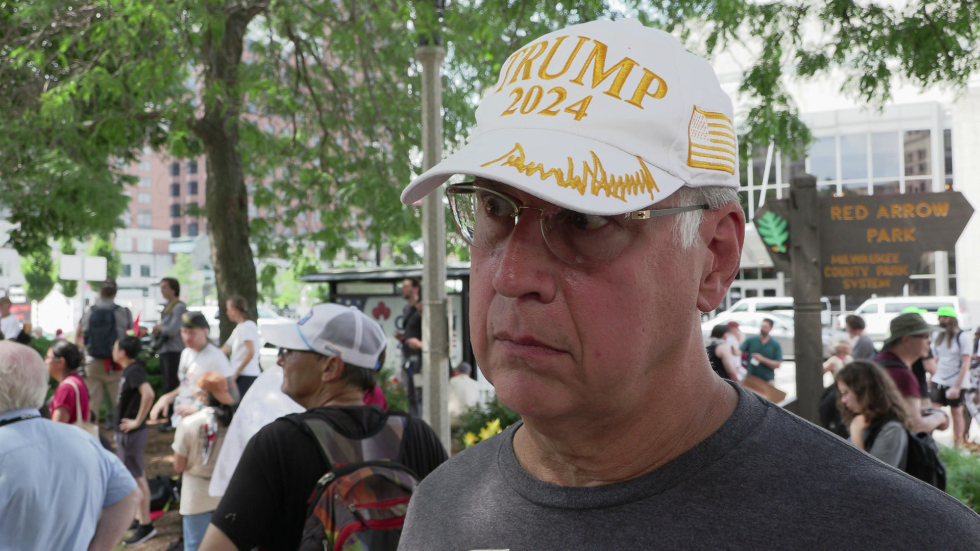 Phillip Reitz speaks while standing outside under a tree with a wood sign reading 'Red Arrow Park" and "Milwaukee County Park System," people and buildings in the background.