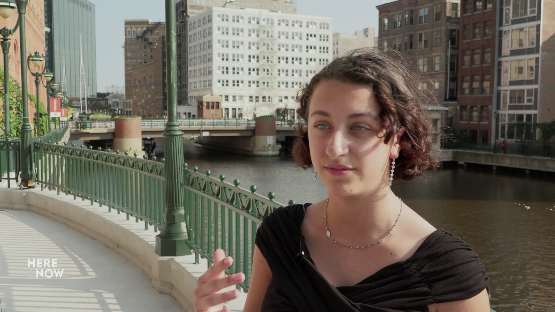 Olivia Kurth gestures with her right hand and speaks while standing on a walkway with a river, buildings and a bridge in the background.