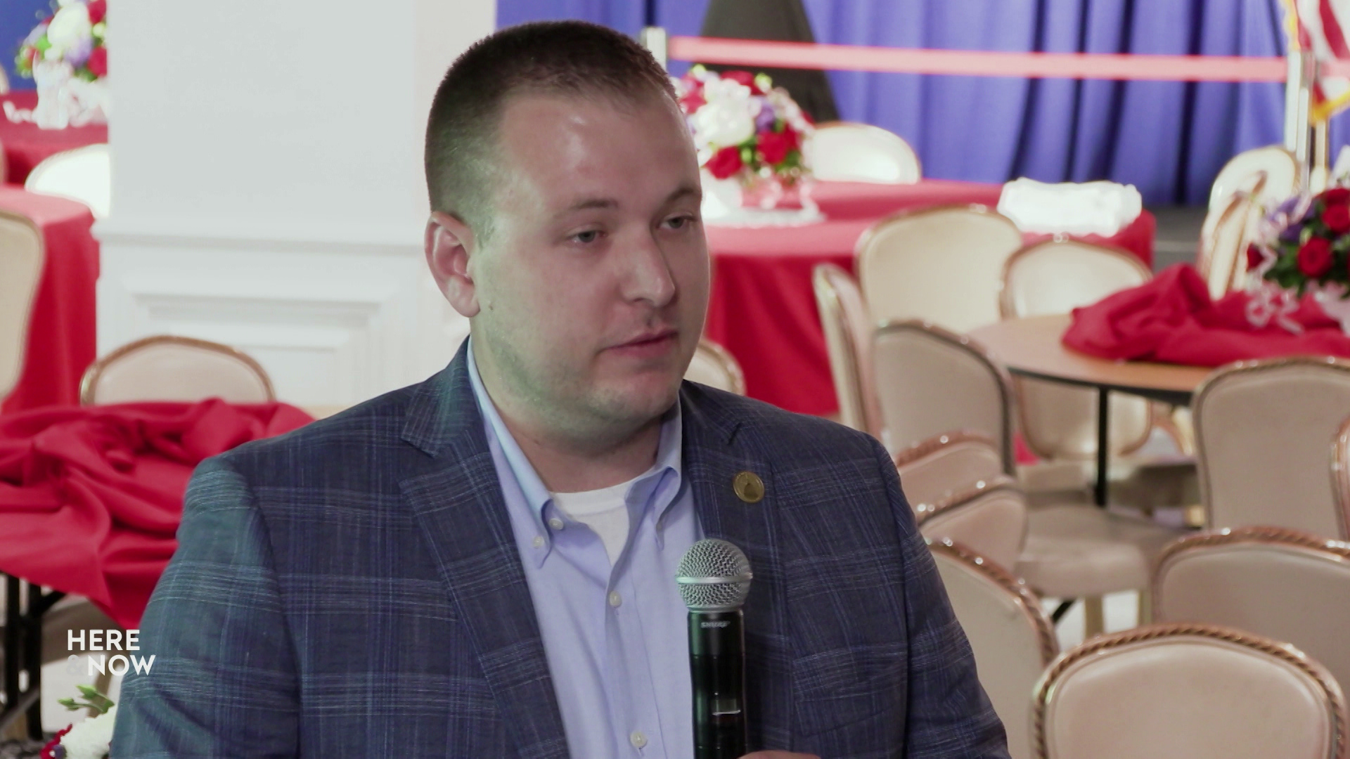 Nik Rettinger speaks into a microphone while sitting in a room with empty chairs and tables with tablecloths and flower centerpieces in the background.