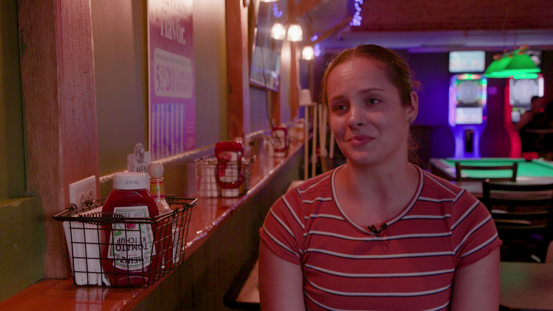 Ariel Malvitt speaks while seated inside with baskets of condiments on shelves with wood paneling and a pool table in the background.