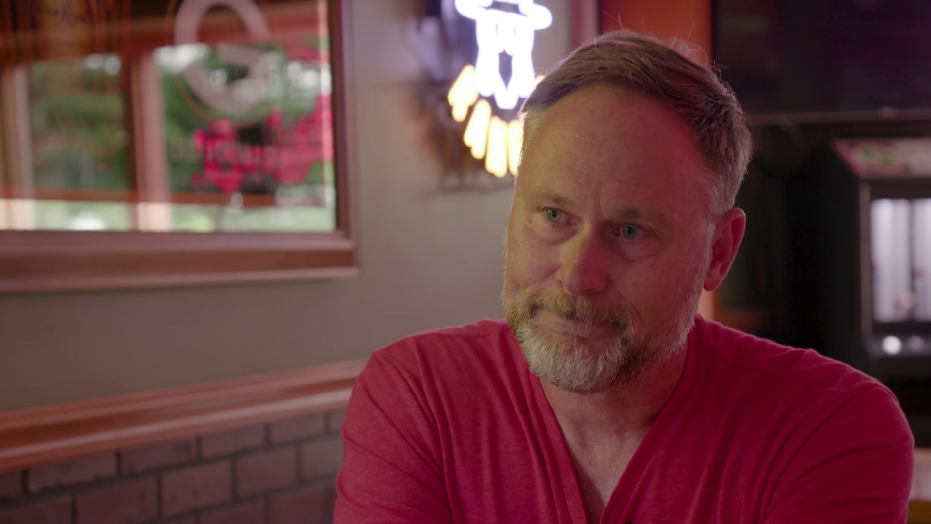 Dominic Roswell speaks while seated inside with wood paneling and a neon sign in the background.