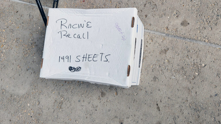 A banker's box with the words Racine Recall and 1491 sheets written in marker on its top sits on a concrete sidewalk.